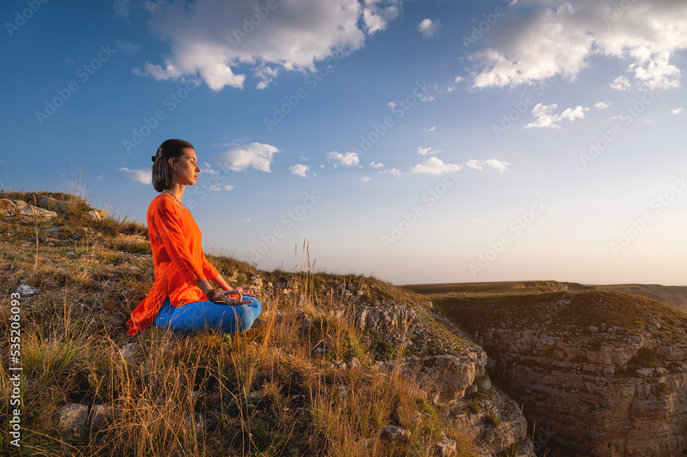 A young slender woman in a lotus position in profile in nature in the mountains sits on the grass, meditates, balances near a rock A young slender woman in a lotus position in profile in nature in the mountains sits on the grass, meditates, balances near a rock