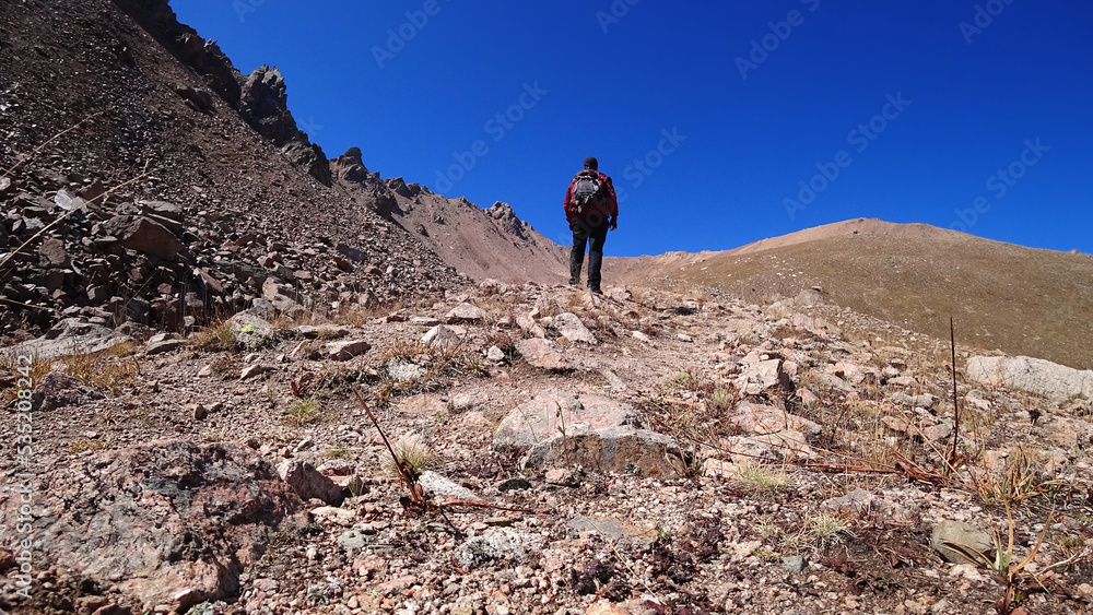 A guy in a tracksuit with a big backpack climbs the peak. Walking along a rocky path. Grass grows among the stones, and in places it is already yellow. Autumn in the mountains. Blue sky rocks.