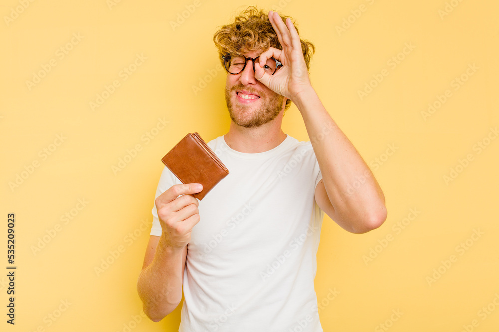 Young caucasian man holding a wallet isolated on yellow background excited keeping ok gesture on eye. Young caucasian man holding a wallet isolated on yellow background excited keeping ok gesture on eye.