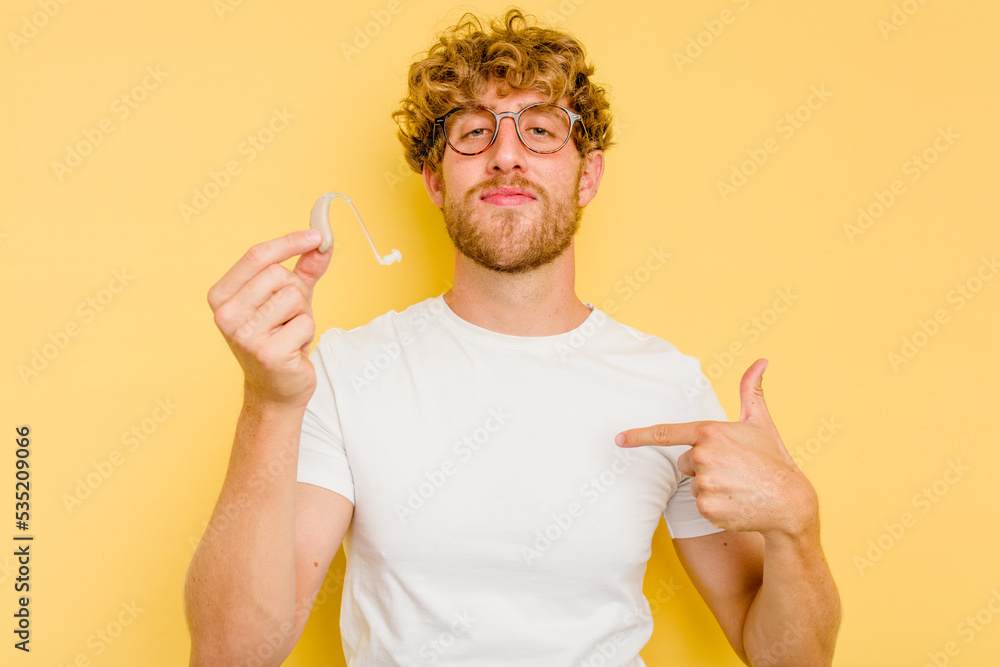 Young caucasian man wearing hearing aid isolated on yellow background person pointing by hand to a shirt copy space, proud and confident Young caucasian man wearing hearing aid isolated on yellow background person pointing by hand to a shirt copy space, proud and confident
