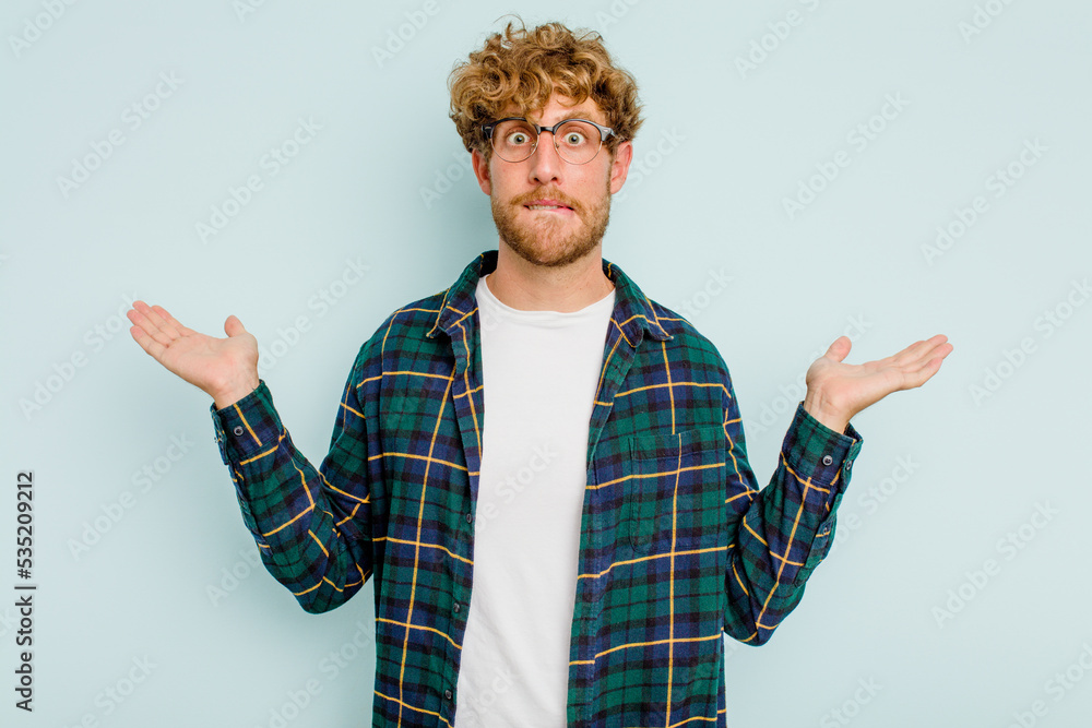 Young caucasian man isolated on blue background confused and doubtful shrugging shoulders to hold a copy space. Young caucasian man isolated on blue background confused and doubtful shrugging shoulders to hold a copy space.