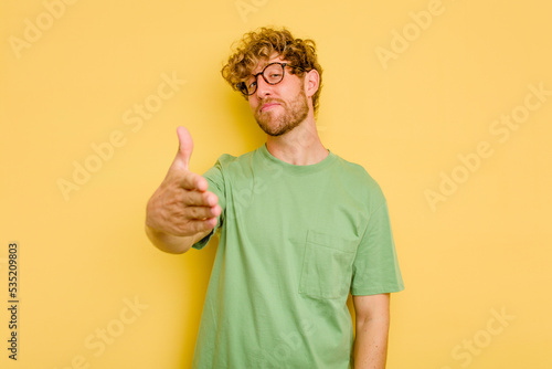 Young caucasian man isolated on yellow background stretching hand at camera in greeting gesture.