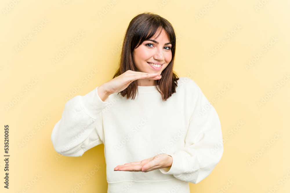 Young caucasian woman isolated on yellow background holding something with both hands, product presentation. Young caucasian woman isolated on yellow background holding something with both hands, product presentation.