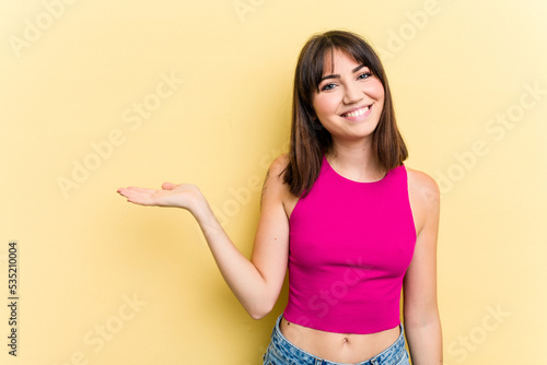 Young caucasian woman isolated on yellow background showing a copy space on a palm and holding another hand on waist.