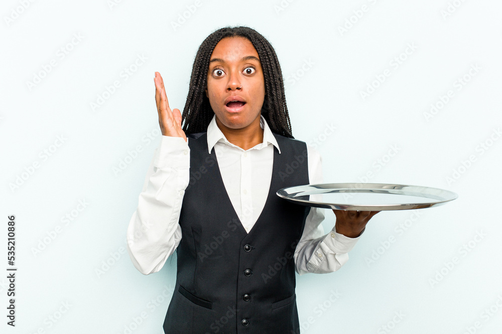 Young African American waitress woman holding a tray isolated on blue background surprised and shocked. Young African American waitress woman holding a tray isolated on blue background surprised and shocked.