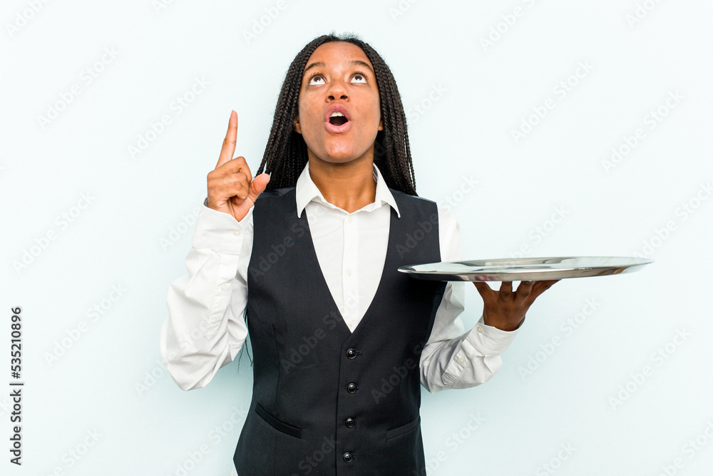 Young African American waitress woman holding a tray isolated on blue background pointing upside with opened mouth. Young African American waitress woman holding a tray isolated on blue background pointing upside with opened mouth.
