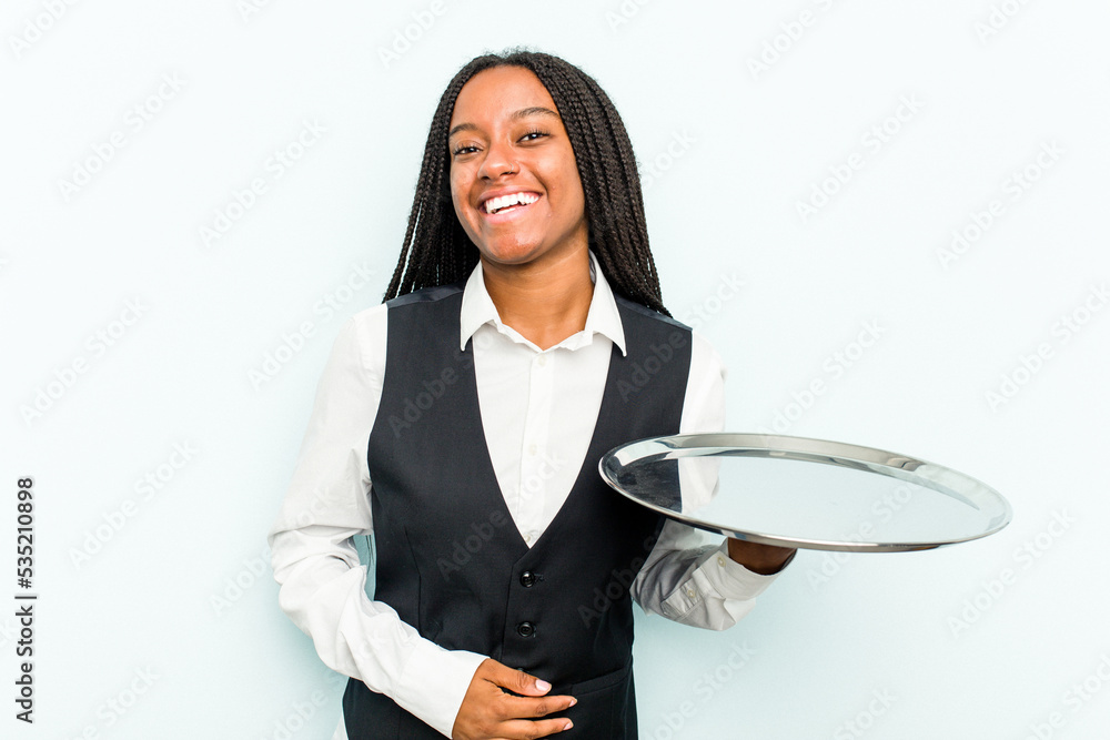 Young African American waitress woman holding a tray isolated on blue background laughing and having fun. Young African American waitress woman holding a tray isolated on blue background laughing and having fun.
