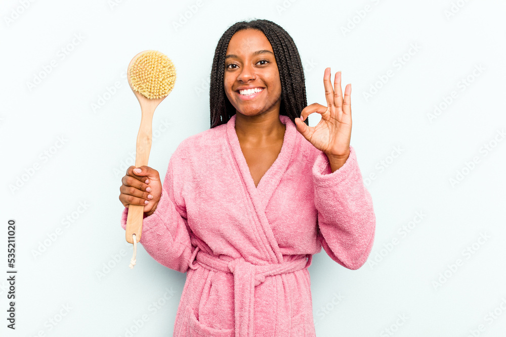 Young African American woman holding a bathtub brush isolated on blue background cheerful and confident showing ok gesture. Young African American woman holding a bathtub brush isolated on blue background cheerful and confident showing ok gesture.