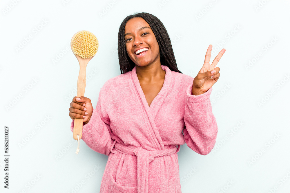 Young African American woman holding a bathtub brush isolated on blue background joyful and carefree showing a peace symbol with fingers. Young African American woman holding a bathtub brush isolated on blue background joyful and carefree showing a peace symbol with fingers.