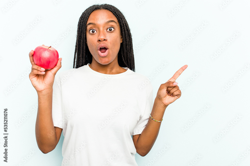 Young African American woman holding an apple isolated on blue background pointing to the side