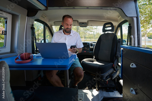 Middle-aged man looking at his cell phone while working on his laptop from his camper van.