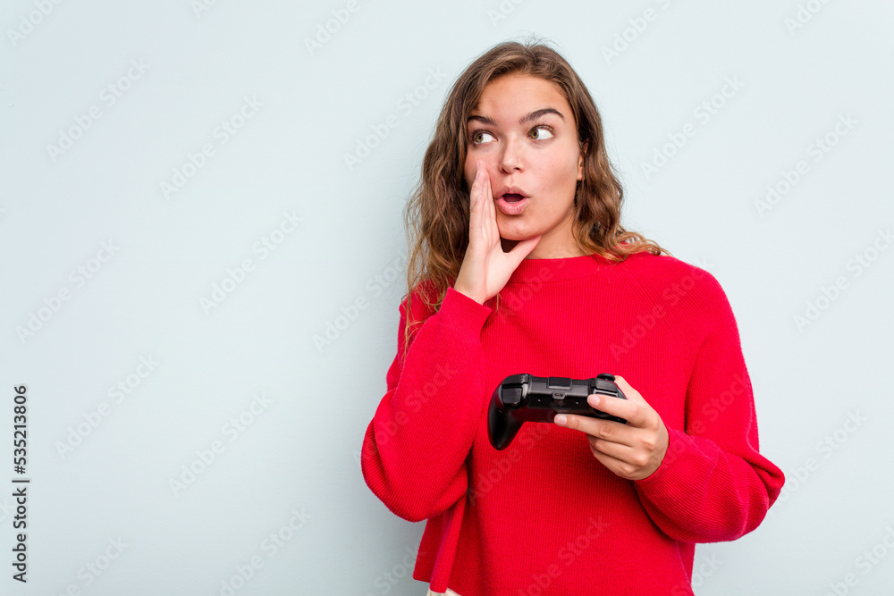 Young caucasian gamer woman holding a game controller isolated on blue background is saying a secret hot braking news and looking aside Young caucasian gamer woman holding a game controller isolated on blue background is saying a secret hot braking news and looking aside