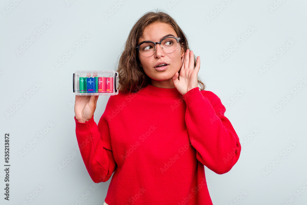 Young caucasian woman holding battery box isolated on blue background trying to listening a gossip. Young caucasian woman holding battery box isolated on blue background trying to listening a gossip.