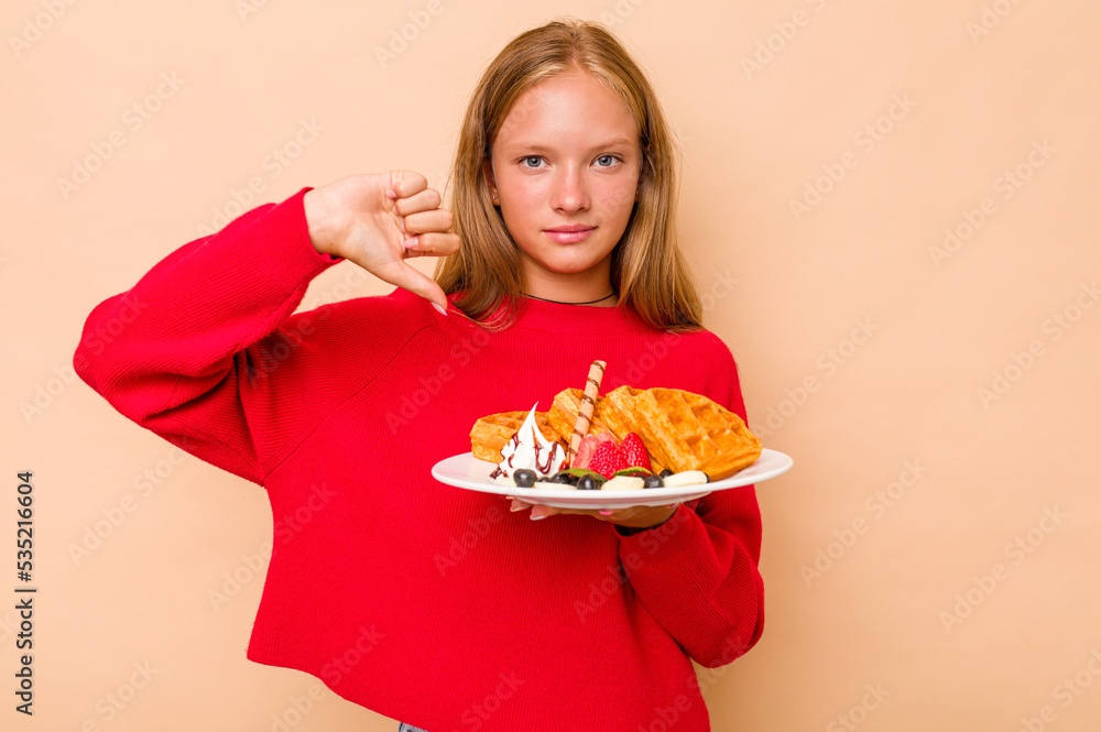 Little caucasian girl holding a waffles isolated on beige background showing a dislike gesture, thumbs down. Disagreement concept.
