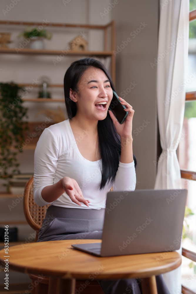 Asian woman sitting at home working on her laptop and talks on the phone comfortably and refreshed on a new day. Asian woman sitting at home working on her laptop and talks on the phone comfortably and refreshed on a new day.