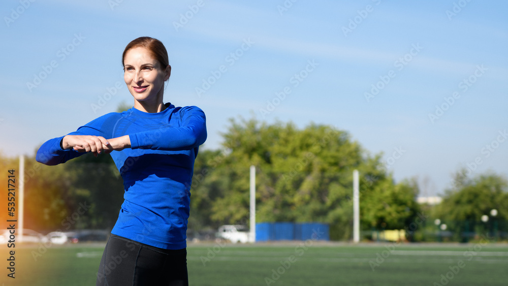 Women and sport. Girl in sportswear does exercises: bends and stretches on the grass at the stadium on a sunny day. Middle aged sportswoman dressed in sportsclothes exercising outdoors Women and sport. Girl in sportswear does exercises: bends and stretches on the grass at the stadium on a sunny day. Middle aged sportswoman dressed in sportsclothes exercising outdoors