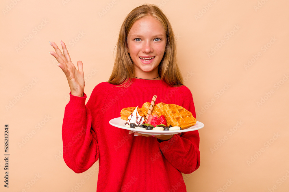 Little caucasian girl holding a waffles isolated on beige background receiving a pleasant surprise, excited and raising hands.