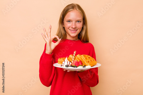 Little caucasian girl holding a waffles isolated on beige background cheerful and confident showing ok gesture.