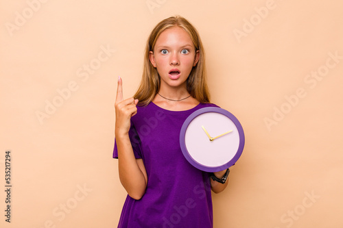 Little caucasian girl holding a clock isolated on beige background having an idea, inspiration concept.