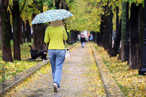 Rain in city, slim woman with umbrella wearing jeans and yellow jacket walking down the street. Rainy weather in autumn park