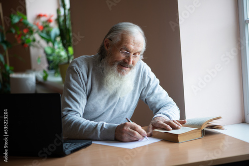 Bearded old man with laptop and book sitting at table and write notes...