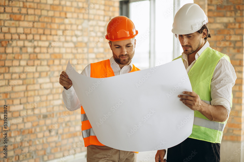 Men architects standing with building plan by the brick wall Men architects standing with building plan by the brick wall