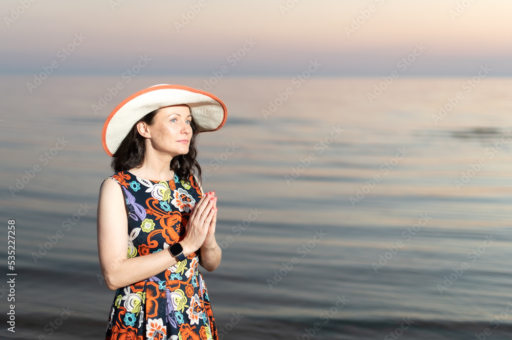a young woman stands with her hands folded in prayer against a sunset sea background