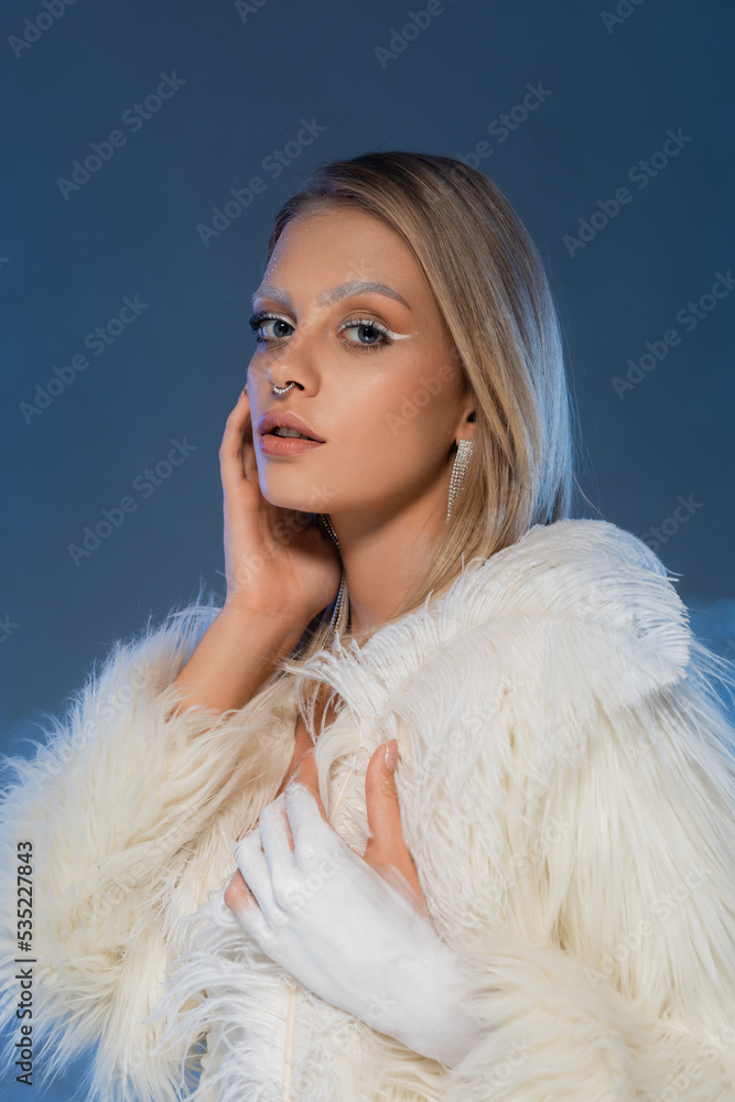 pierced woman with winter makeup posing in white faux fur jacket on dark blue. pierced woman with winter makeup posing in white faux fur jacket on dark blue.