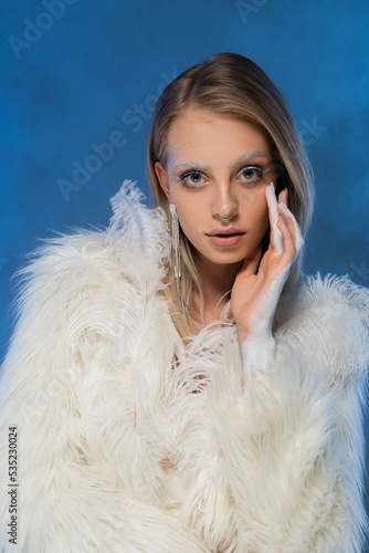 pierced young woman with winter makeup posing in white faux fur jacket and looking at camera on dark blue.