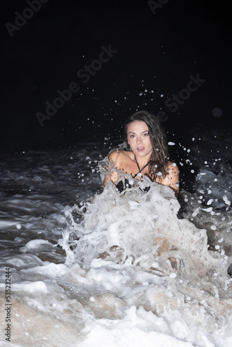 Fun fit young beauty female model kneeling in the sea while the waves are splashing at night