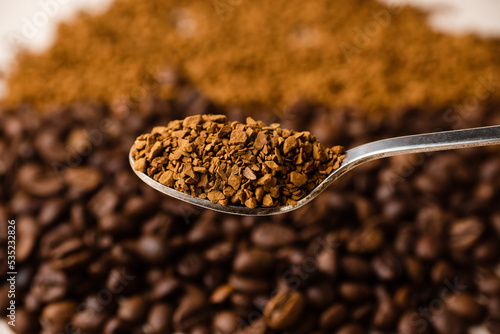 A spoonful of instant coffee against the background of a cluster of coffee beans and a scattering of instant coffee.