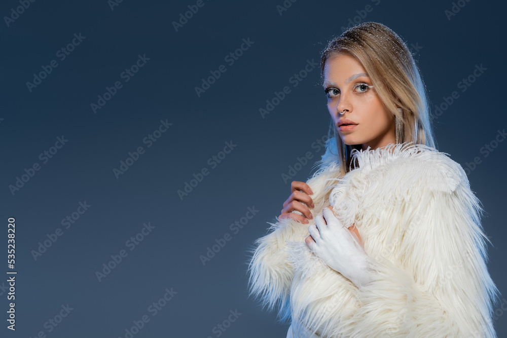 blonde woman with piecing posing in white faux fur jacket and holding feather on dark blue background. blonde woman with piecing posing in white faux fur jacket and holding feather on dark blue background.