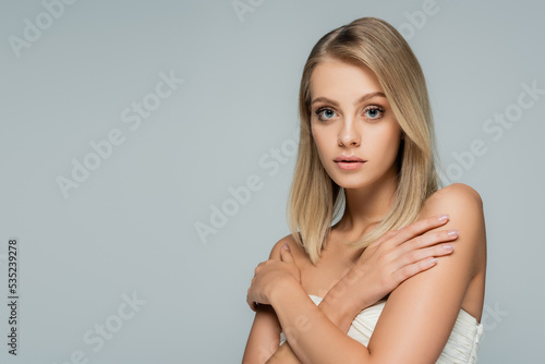young blonde woman with bare shoulders and natural makeup posing with crossed arms isolated on grey.