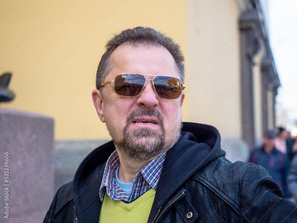 Very angry and aggressive man with beard in dark jacket and sunglasses walking in old town Very angry and aggressive man with beard in dark jacket and sunglasses walking in old town