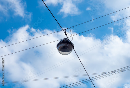 Black round glass pendant lamp hanging in the at the crossing of black electrical wires over city road against the blue sky