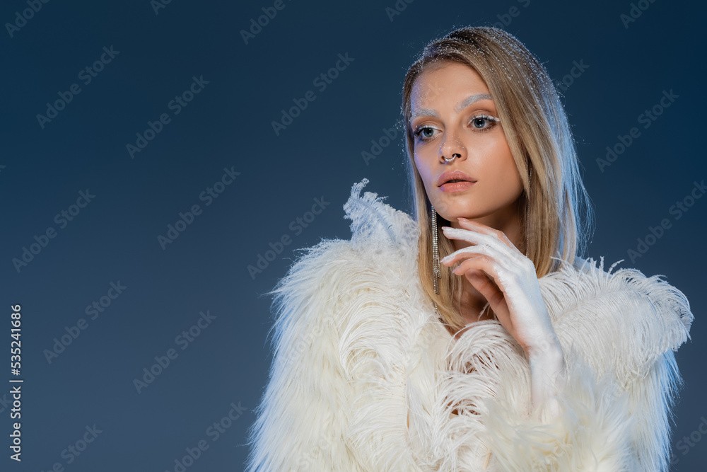 blonde woman with piecing posing in white faux fur jacket with feathers on dark blue background. blonde woman with piecing posing in white faux fur jacket with feathers on dark blue background.
