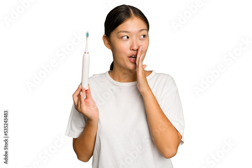 Young Asian woman holding an electric toothbrush isolated on green chroma background is saying a secret hot braking news and looking aside