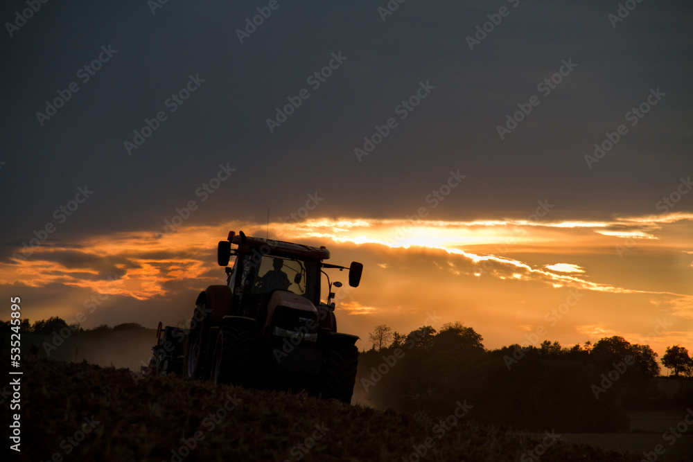 Fototapeta premium Tractor harrowing reddish brown, dusty, clayey soil in a hilly landscape at sunset