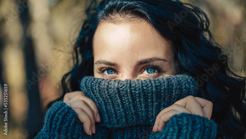 A pretty fashionable young woman with natural makeup and beautiful blue eyes covers her lips with a sweater outdoors. A fashionable girl is resting on the orange autumn foliage in the forest.