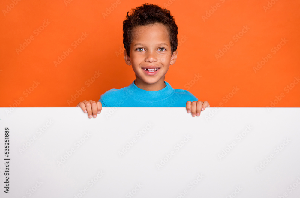 Photo portrait of charming little boy standing behind white banner empty space wear trendy blue clothes isolated on orange color background Photo portrait of charming little boy standing behind white banner empty space wear trendy blue clothes isolated on orange color background