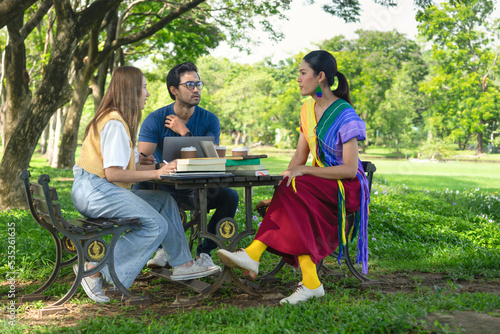 group of friends studying outdoor in the park on weekend, selective focus at a man, young asian people chatting together, there are device for learning on table such as laptop,the books,coffee cups