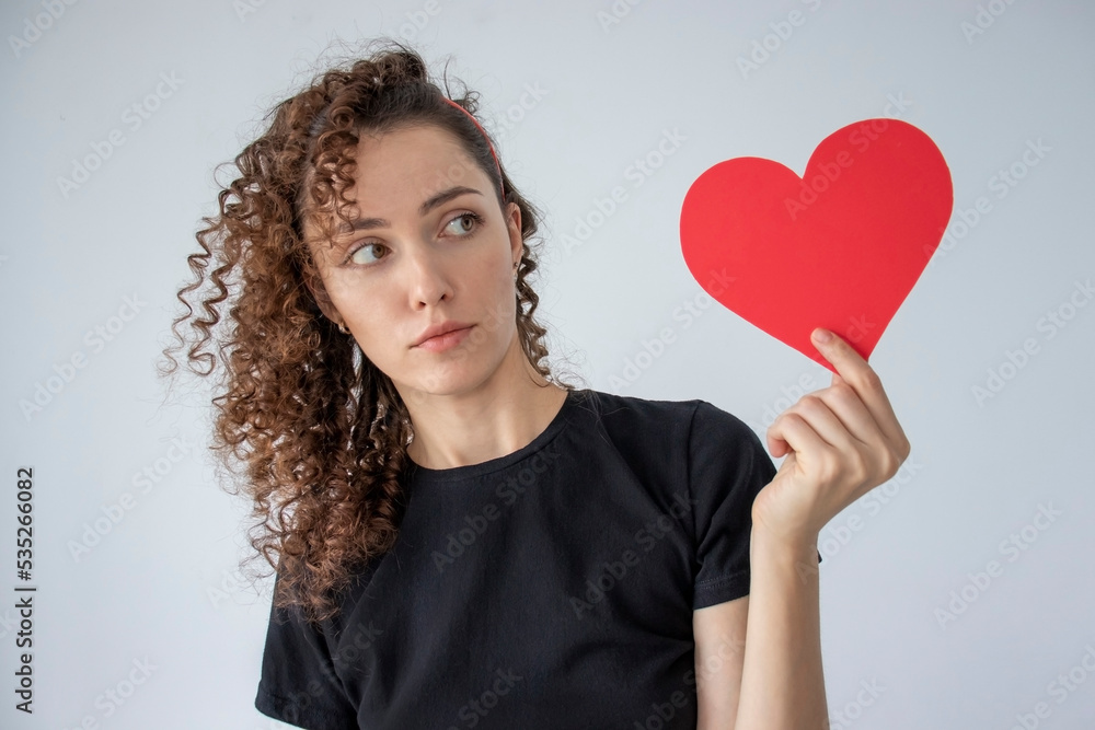 Portrait of an attractive curly-haired girl holding a large red paper heart with a surprised face on a gray background Portrait of an attractive curly-haired girl holding a large red paper heart with a surprised face on a gray background