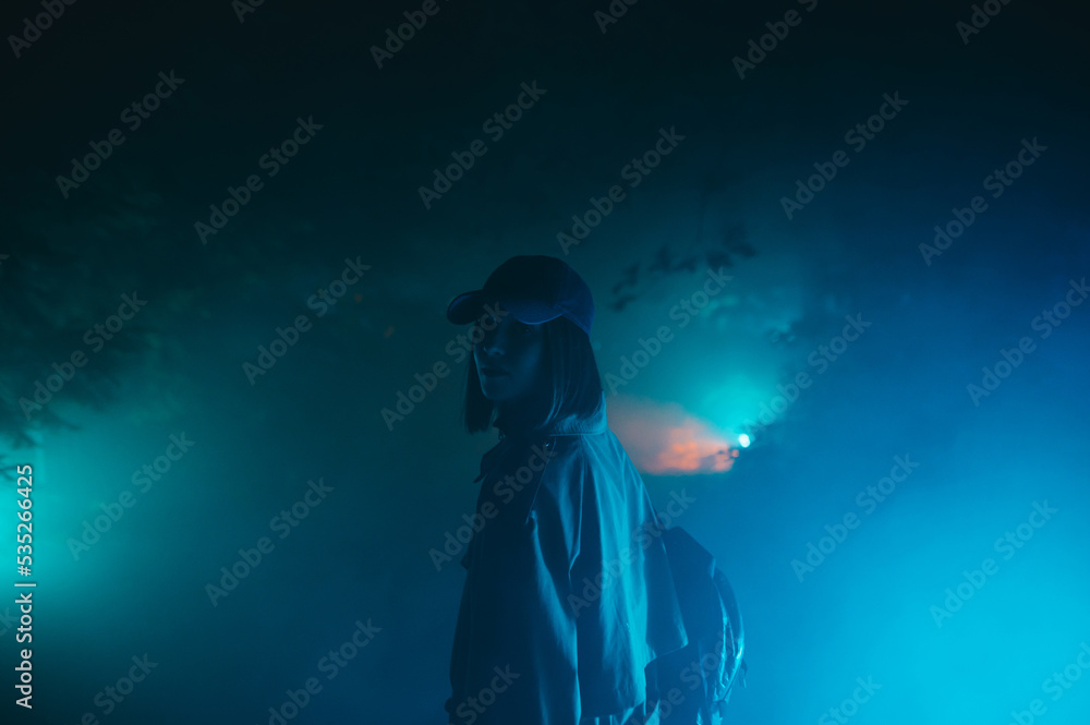 Attractive woman in casual clothes stands in the fog in the park in blue light and looks at the camera with a serious face.