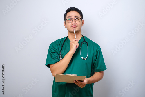 Thoughtful smart asian nurse in scrubs looking away and thinking, while holding clipboard and putting pen on his chin.