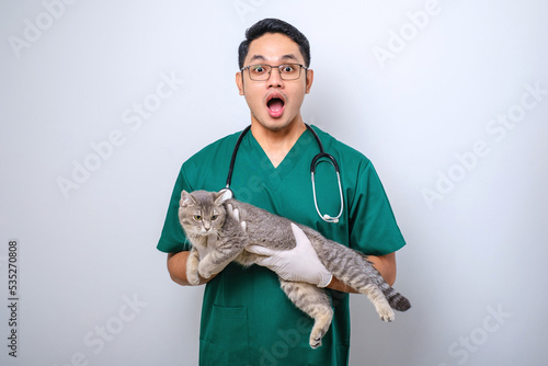Shocked doctor in vet clinic examining cat with stethoscope, gasping amazed while holding cat in his arm
