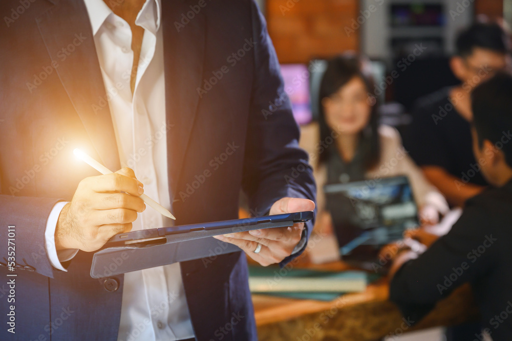 Obraz premium male business man holding a tablet meeting about the project standing at an office desk with a group of colleagues in the background working on a laptop computer
