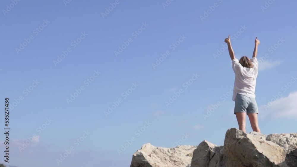 View from the back. A female tourist raises her hands up standing on the edge of a cliff against the sky. View from the back. A female tourist raises her hands up standing on the edge of a cliff against the sky.