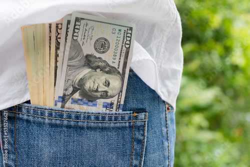 Dollar banknotes in the back pocket of jeans on background as a public park. Finance and tax concept. 