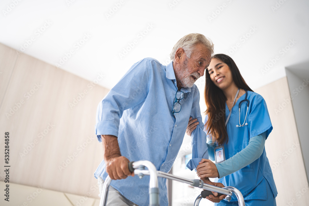 Fototapeta premium Portrait of Asian young nurse helping old elderly disable man grandfather to walk by using walker equipment in the bedroom. Senior patient of nursing home moving with walking frame and nurse support