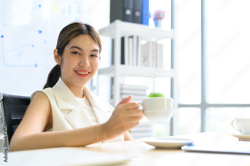 Cheerful businesswoman drinking coffee while sitting at table during meeting in light office. Cheerful businesswoman drinking coffee while sitting at table during meeting in light office.